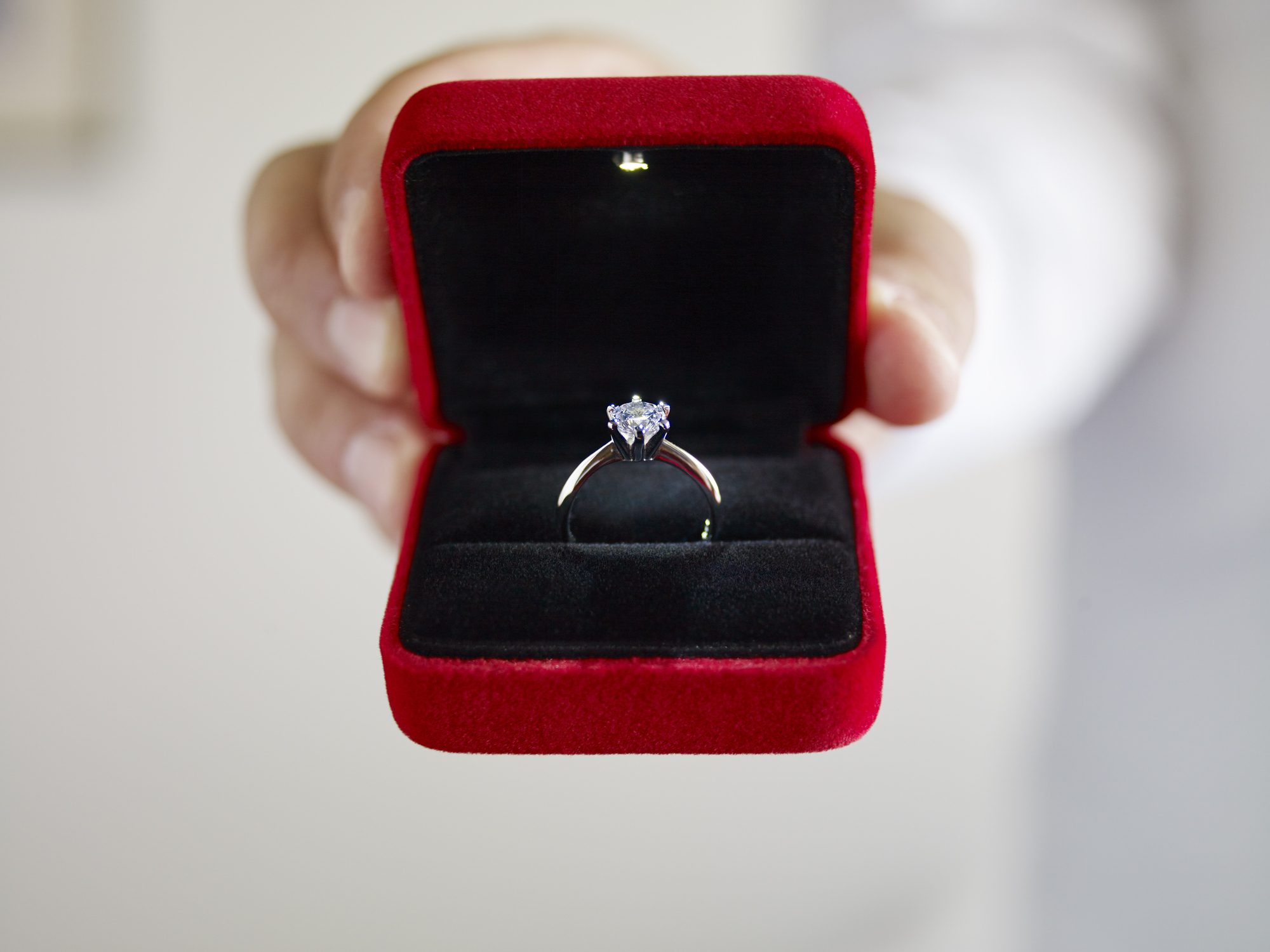 A person's hand holds out a red velvet ring box with a diamond solitaire engagement ring inside