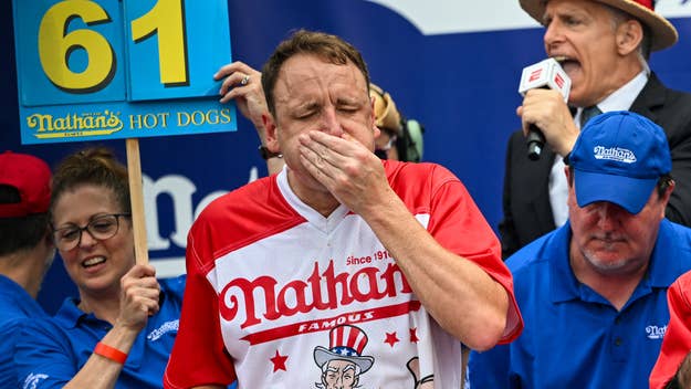 Joey Chestnut and others at Nathan's Famous Hot Dog Eating Contest. Joey holds his mouth, a sign reading "61" is behind him, and an announcer speaks into a microphone
