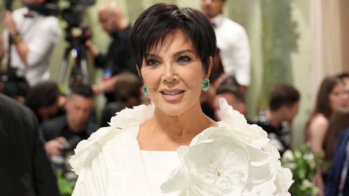 Kris Jenner smiling at an event, wearing a white outfit adorned with a large floral accessory