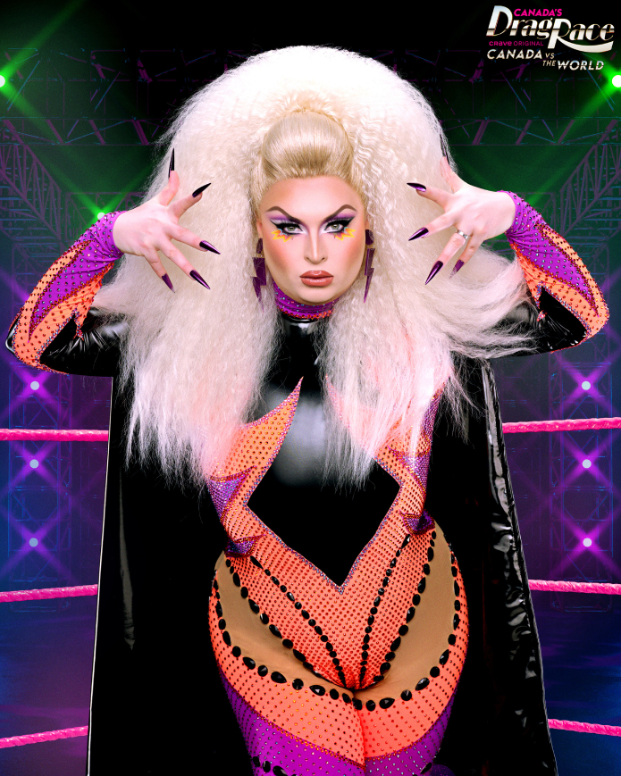 Drag queen in elaborate stage outfit with large curly hair and striking makeup, posing with styled nails in front of a "Canada's Drag Race: Canada vs. The World" backdrop. Names unknown