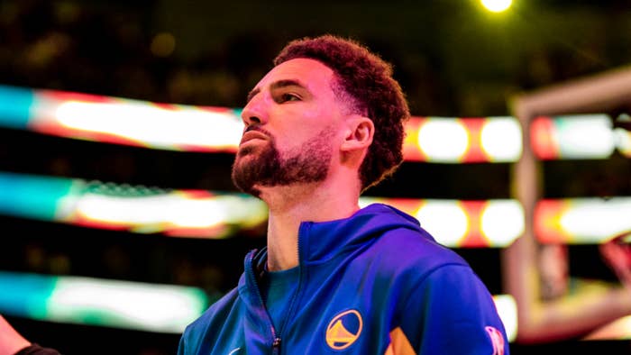 Klay Thompson stands on a basketball court in a blue and yellow Golden State Warriors jacket, looking focused during a game