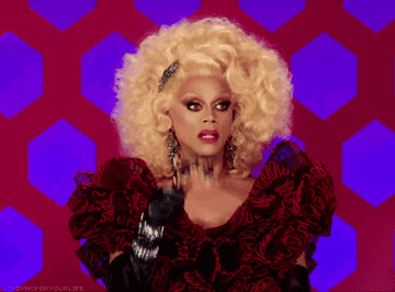 RuPaul with voluminous blonde hair, wearing an elaborate red and black outfit, making a surprised expression on a patterned backdrop