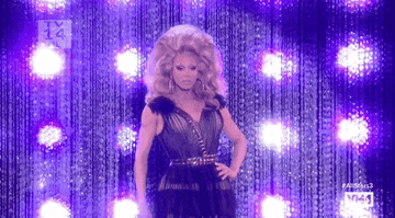 RuPaul on stage in a glamorous, sequined dress with a plunging neckline and voluminous hairstyle, standing in front of a glittering backdrop with bright lights. #AllStars3
