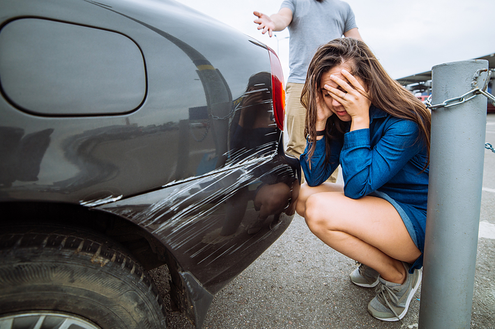 駐車場で車の傷を確認する男性と顔を覆う女性。車の後部に目立つ傷がある。