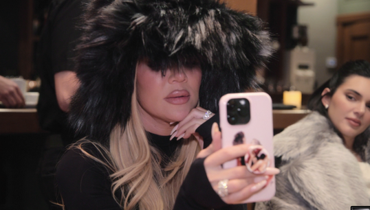 Khloé Kardashian is taking a selfie while wearing a large, fluffy hat. Kendall Jenner sits behind her, also wearing a fur coat