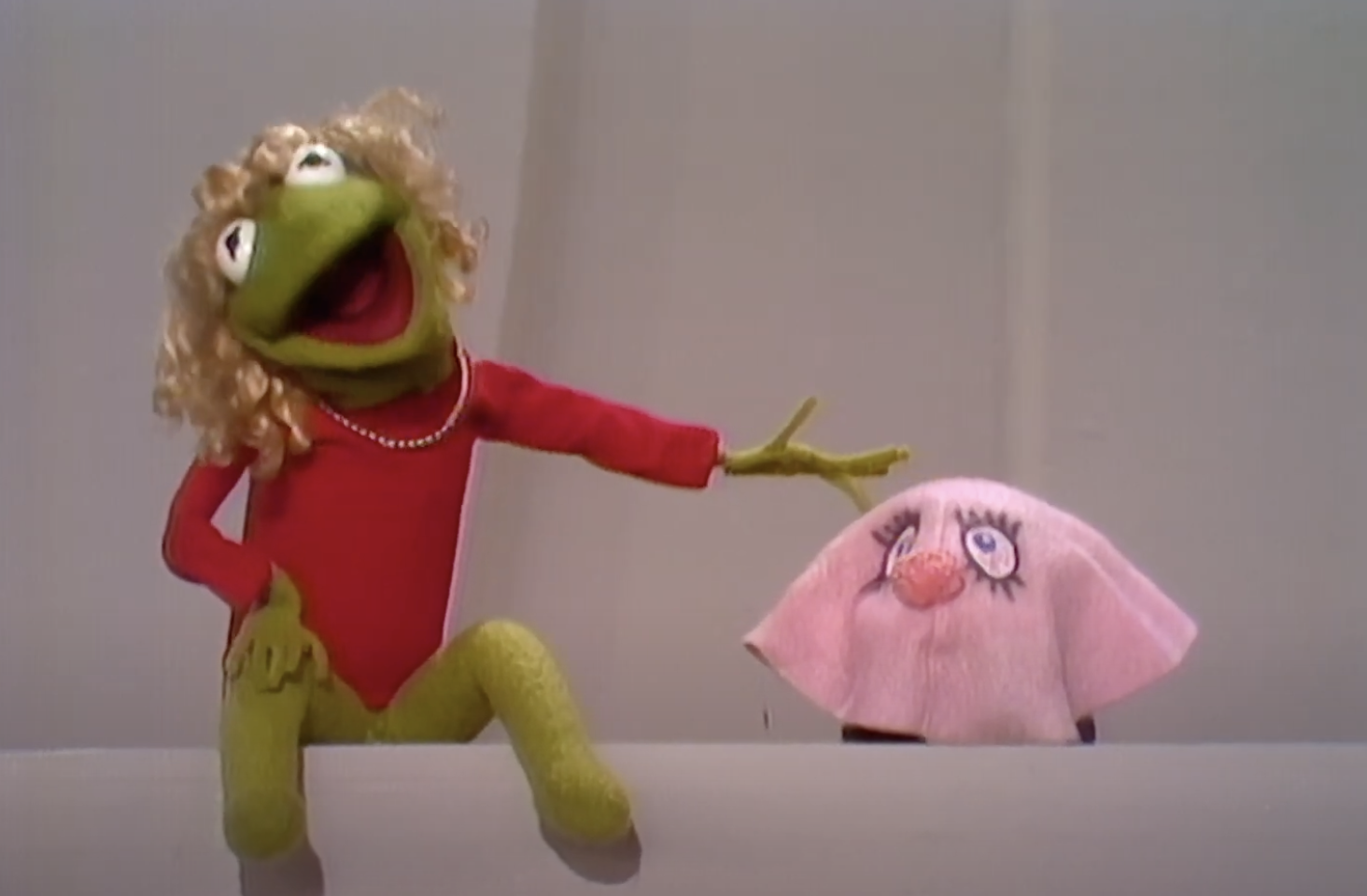 Kermit the Frog in a red leotard and blonde wig, extends his arm towards Miss Mousey, a pink puppet with large eyes and a small red nose