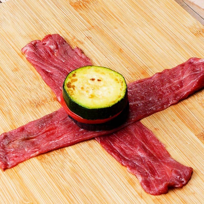 木製ボードに十字に置かれた生肉の上に、なすとズッキーニとトマトのスライスが重ねられた料理。