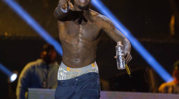 Rapper Kodak Black performs shirtless on stage, holding a water bottle and pointing towards the camera; background features bright, dynamic stage lights