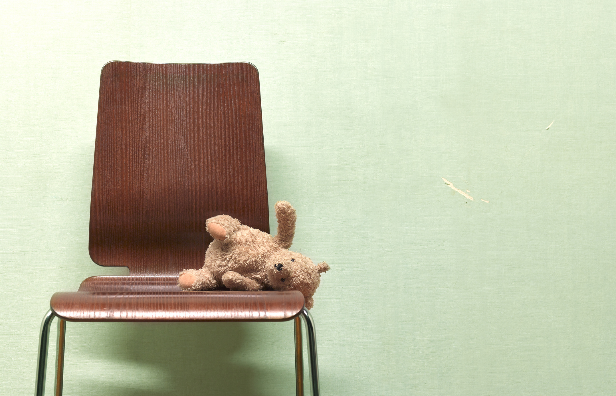 A small, plush teddy bear lies on a wooden chair against a plain wall