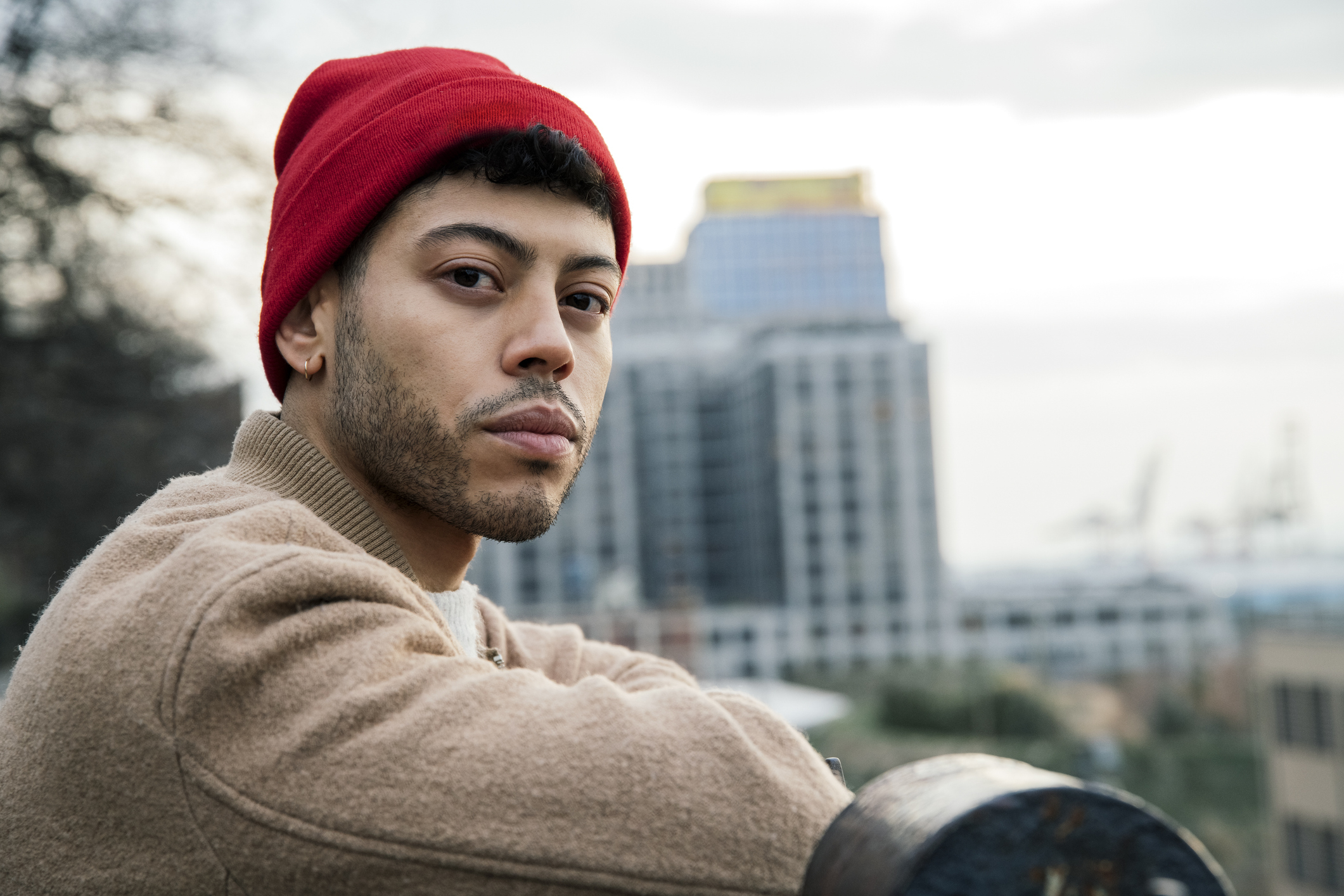A man wearing a red beanie and a tan jacket looks intently at the camera with a cityscape in the background