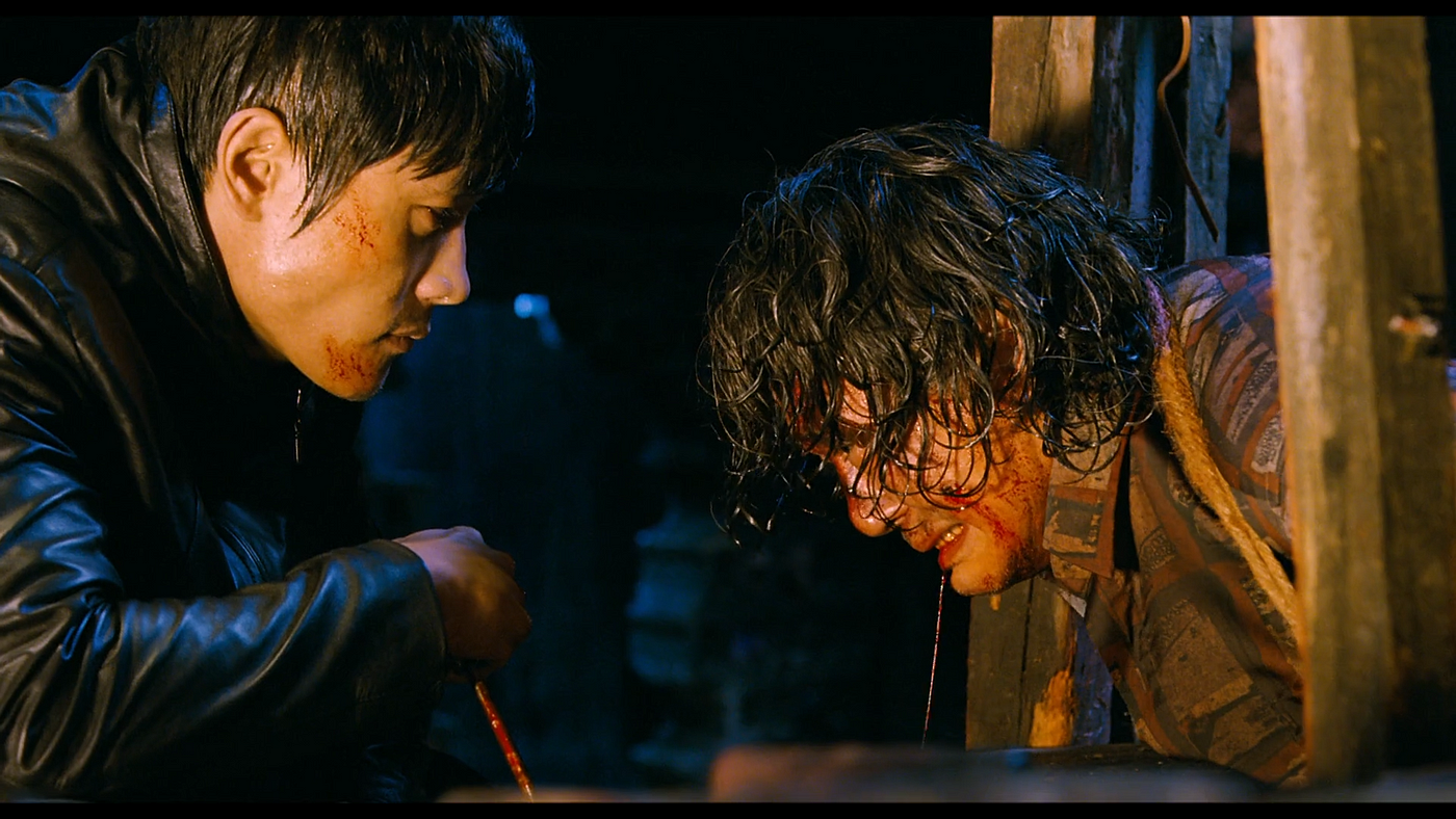 Two individuals are in a dark scene; one person with blood on his face stares at the other person, who is restrained and bloodied. Names unknown