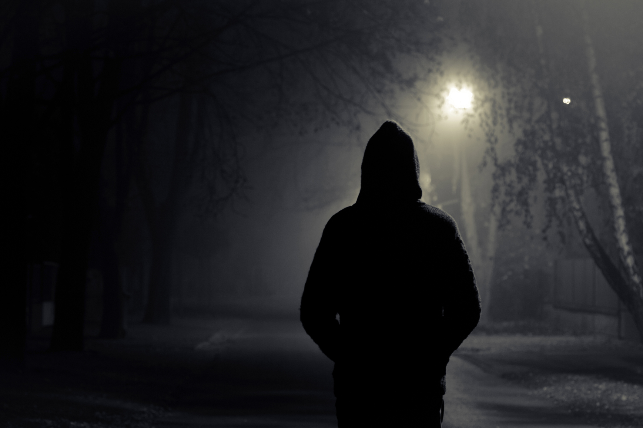 A person in a hooded jacket is standing alone on a foggy, dimly lit street at night