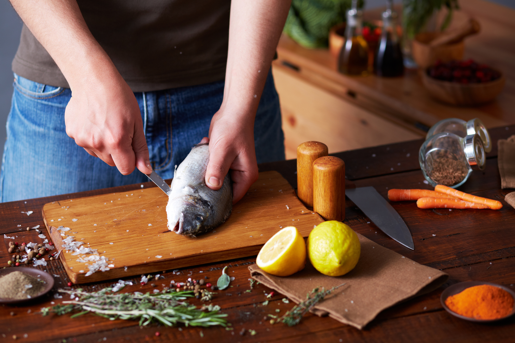 A person is shown gutting a fish on a wooden cutting board, surrounded by various kitchen ingredients like spices, vegetables, and lemon