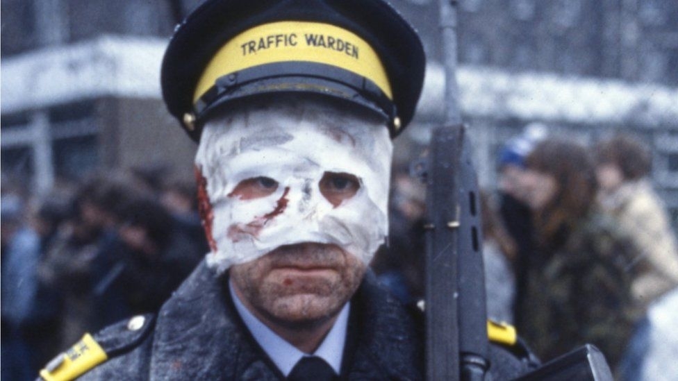 Man in a traffic warden uniform with a bandaged face and holding a rifle, standing outdoors with a crowd in the background