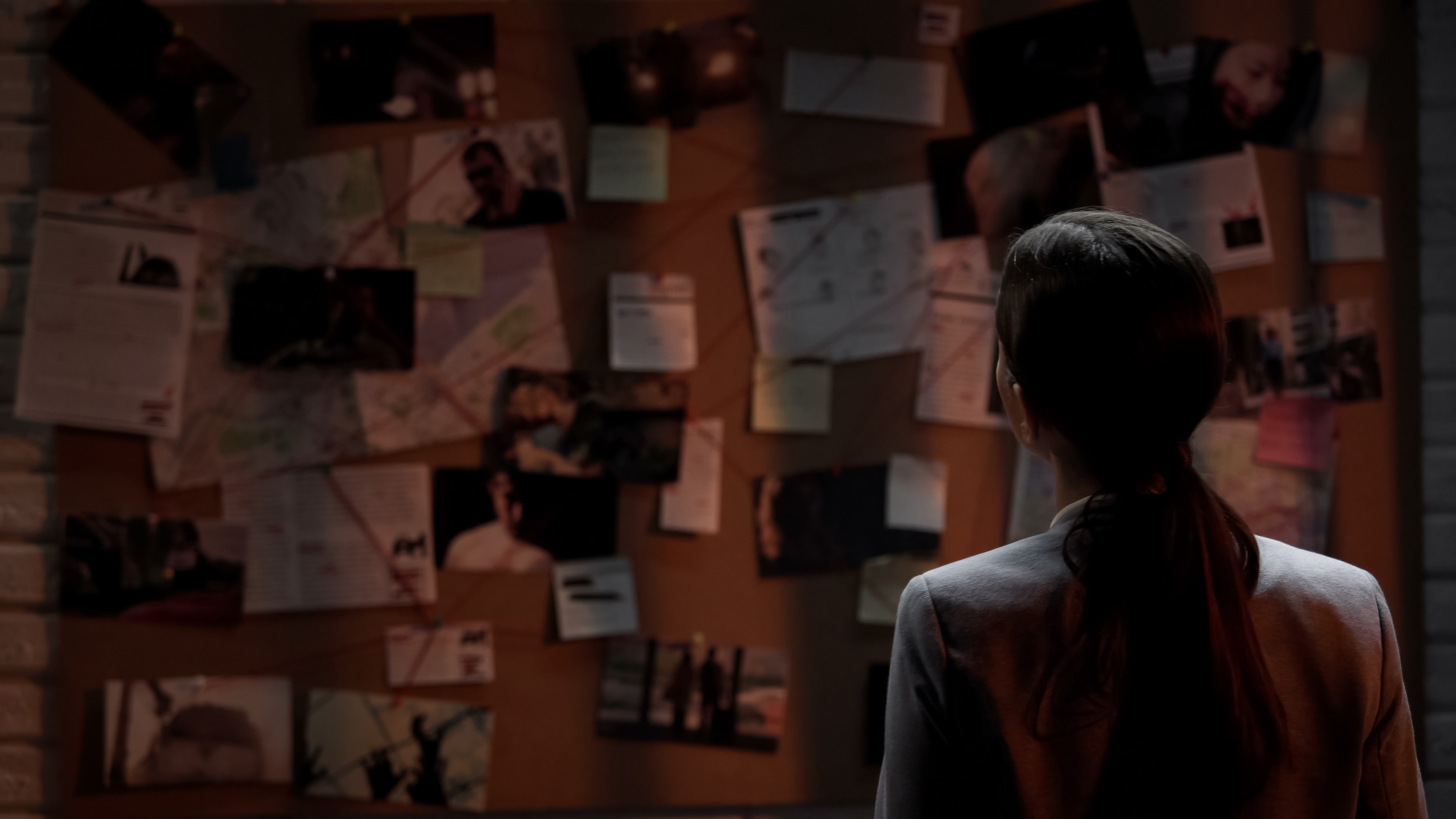A person with a ponytail looks at a bulletin board covered with photos, maps, and documents