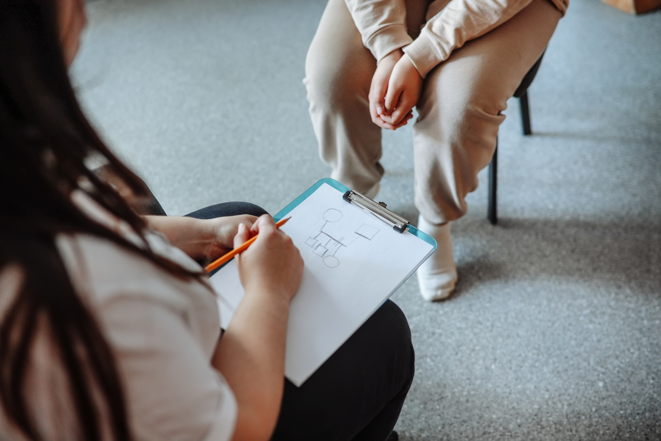A person holds a clipboard drawing a family tree while another person sits beside them with hands clasped. Faces are not visible