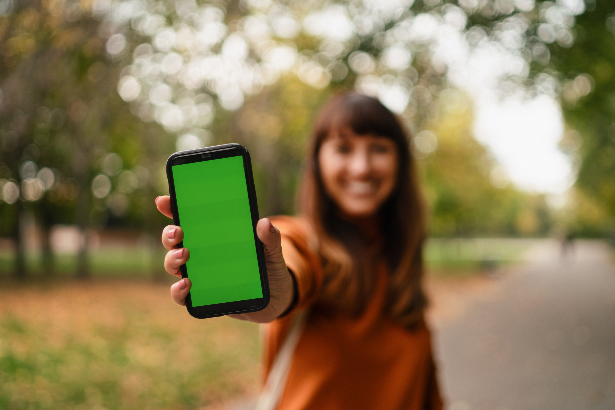 A person holding a smartphone with a green screen toward the camera, standing on an outdoor pathway with trees in the background
