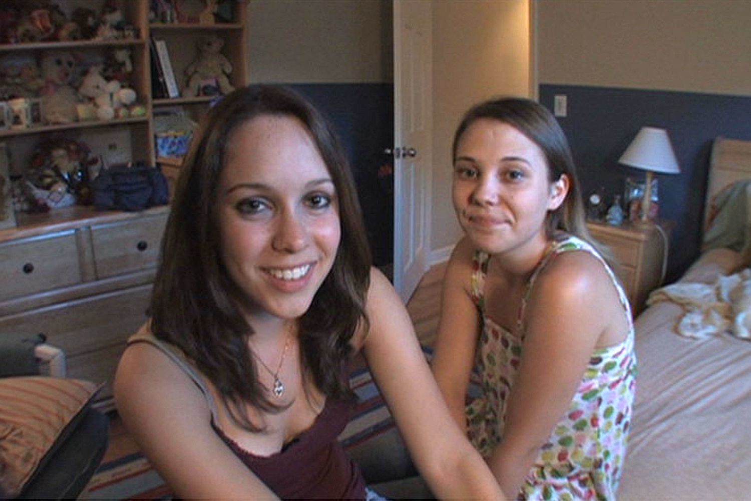 Two women, both smiling, are sitting in a bedroom. One is wearing a tank top, and the other is wearing a polka-dot dress. The bedroom has a cozy and casual appearance