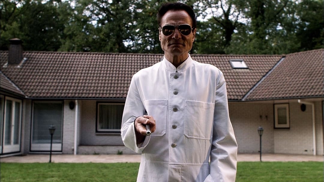 A person in a white lab coat stands outside a house, pointing a remote control toward the camera, with a stern expression and sunglasses on