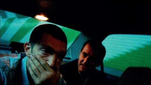 Two men are inside a car. The man in the foreground looks contemplative with his hand covering part of his face, while the man in the background speaks to him