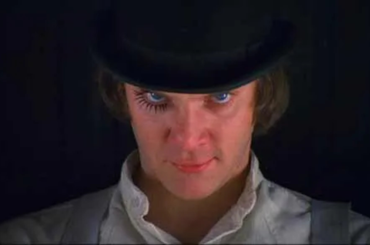 A person with intense blue eyes, wearing a black bowler hat, white shirt, and long fake eyelashes on one eye, appearing as a character from "A Clockwork Orange."