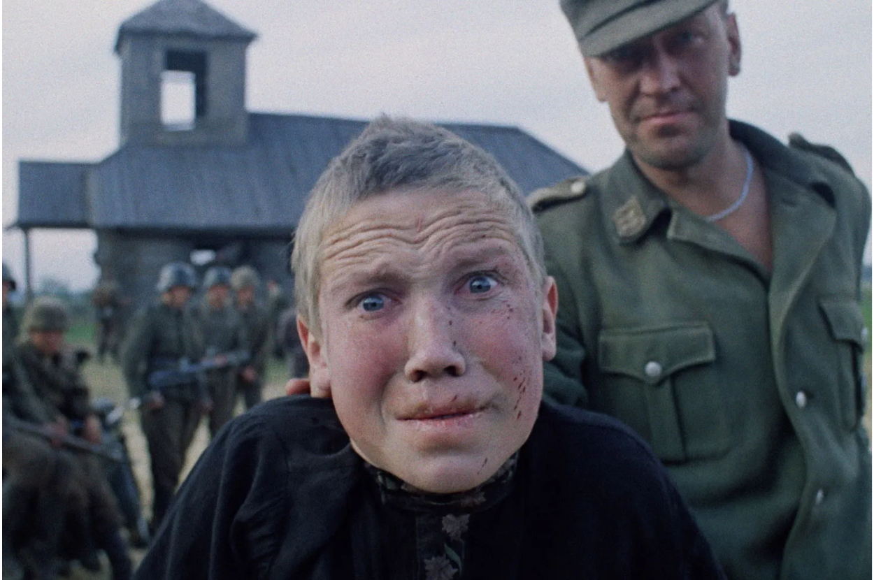 Scene featuring a distressed boy with short hair being held by a soldier in uniform. A building and other soldiers are in the background