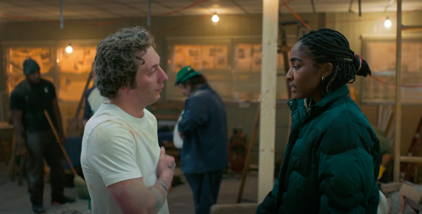 Jeremy Allen White and Ayo Edebiri stand in a busy workshop, engaged in a serious conversation. People are working in the background
