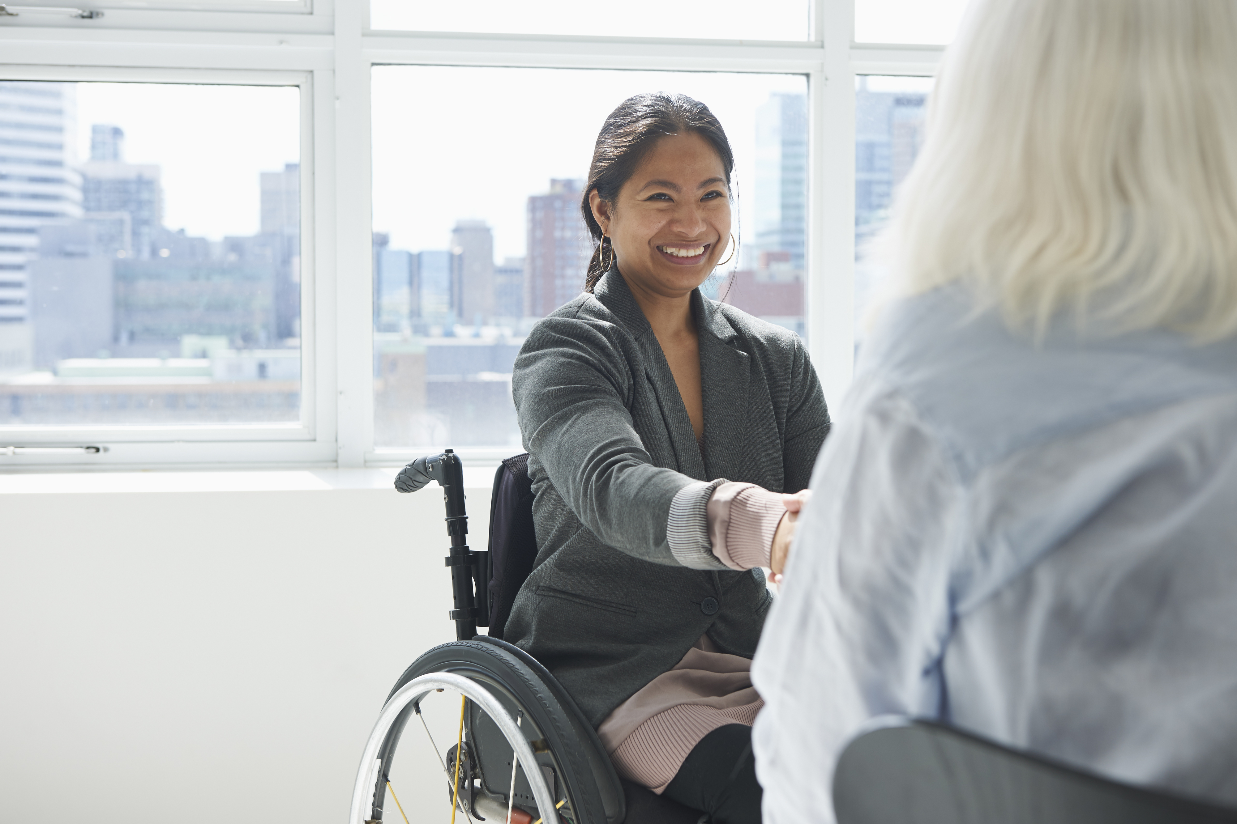A businesswoman in a wheelchair shakes hands with another person in front of a window with a city skyline view