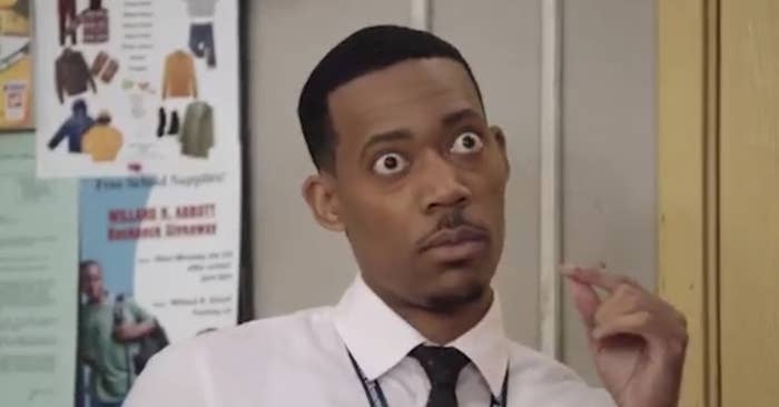 A man in a white dress shirt and black tie, with an expressive look on his face, is standing in front of a bulletin board covered with various flyers and notices