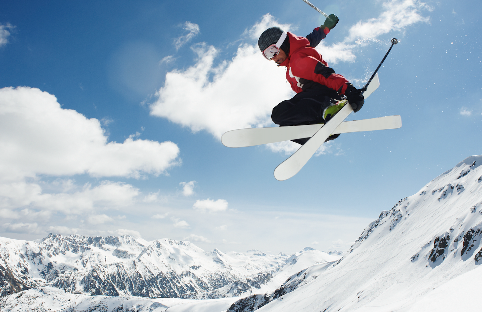 A skier in mid-air performs a stylish jump over a snow-covered mountain range with a clear sky backdrop