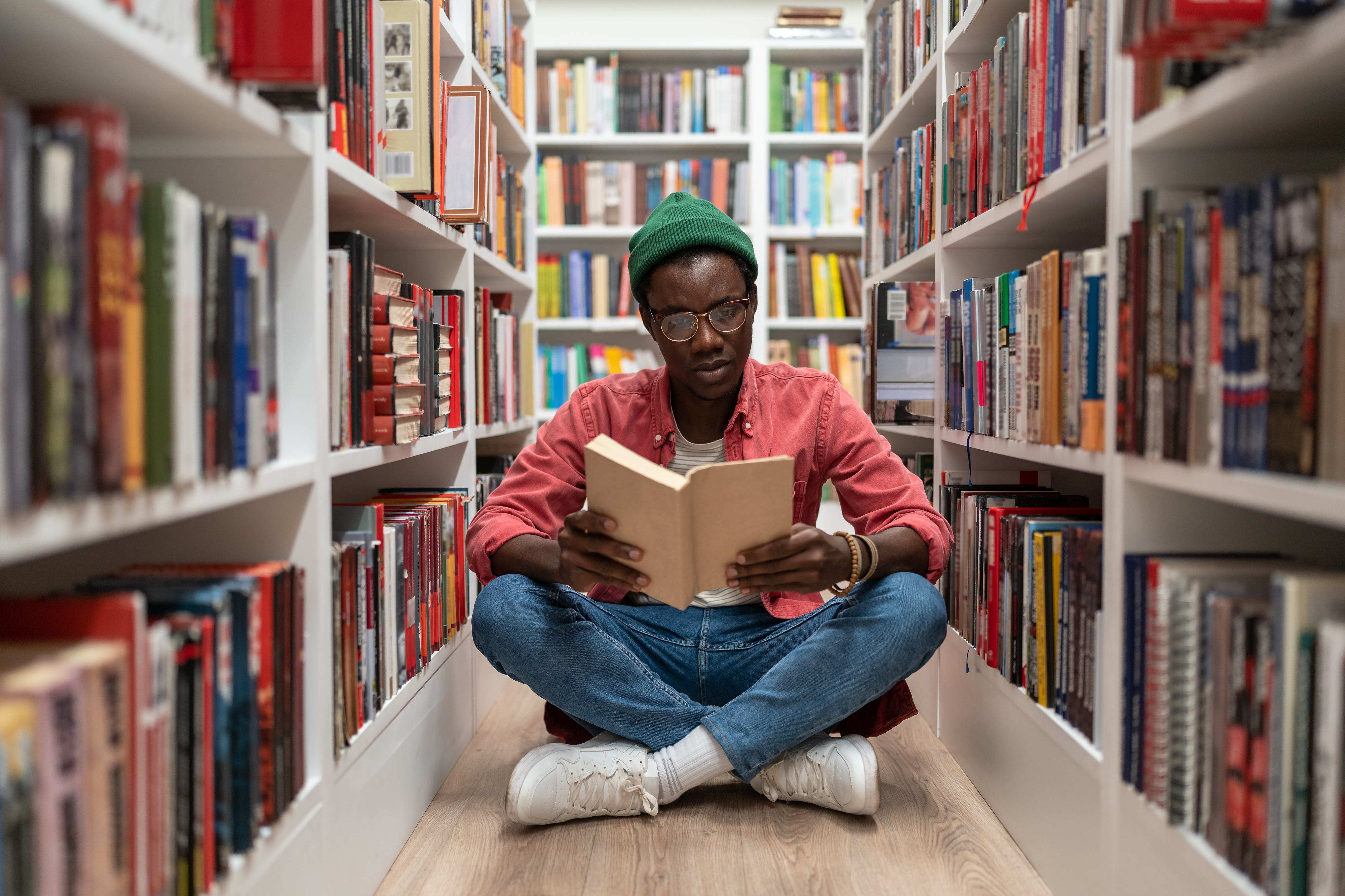 A person sits cross-legged on the floor between bookshelves in a library, engrossed in reading a book