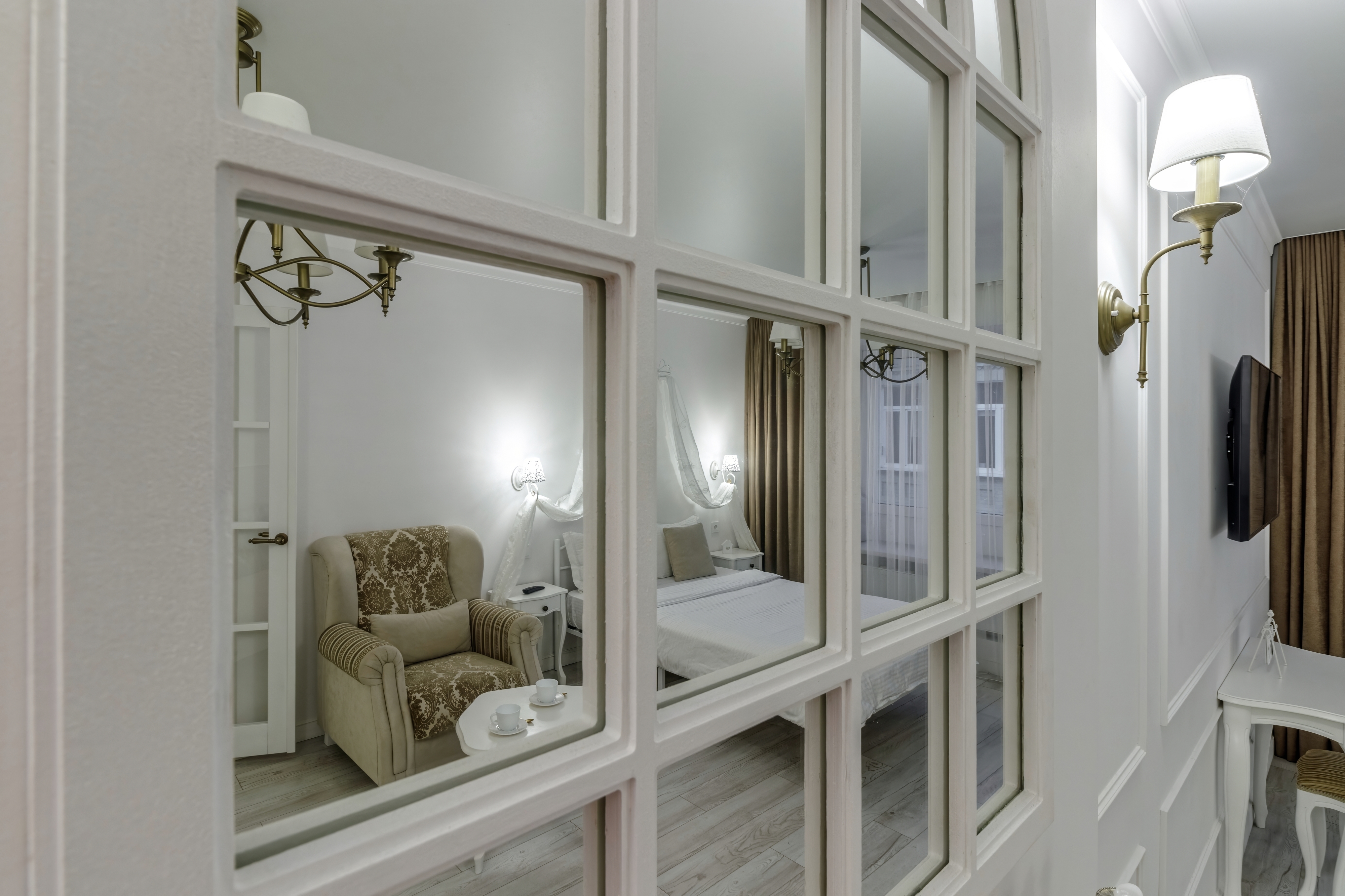 Elegant boudoir view featuring mirrored paneling, a cozy patterned armchair, a small table with a teapot set, and a well-lit bedroom with draped bed