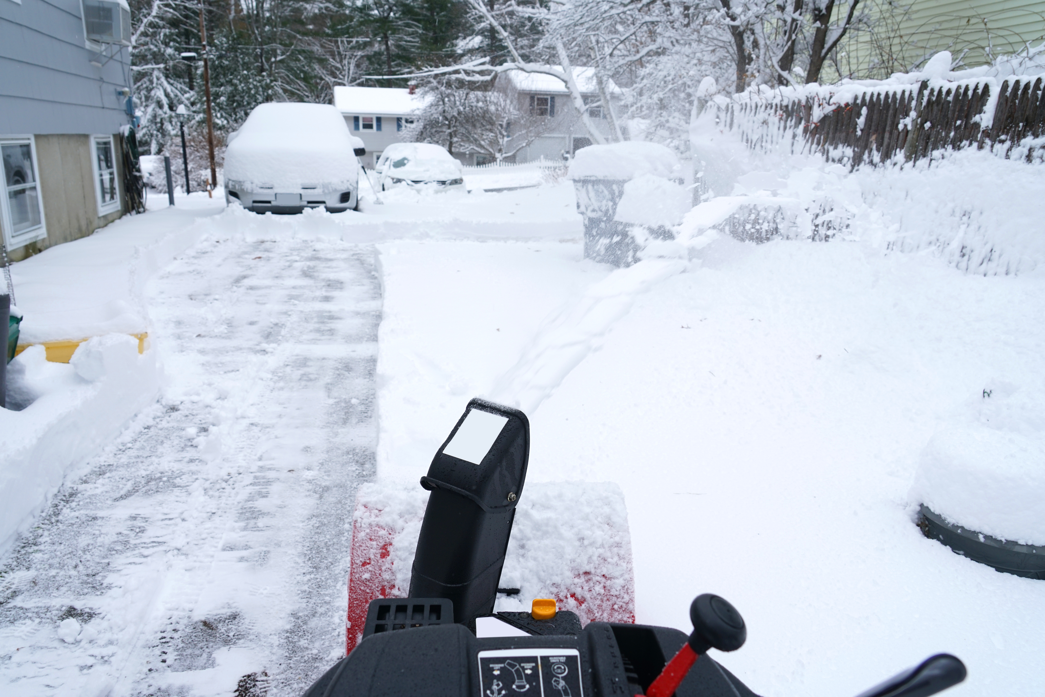 Snow-covered driveway and yard with a snow blower clearing a path. Snow-covered cars and fences visible in the background