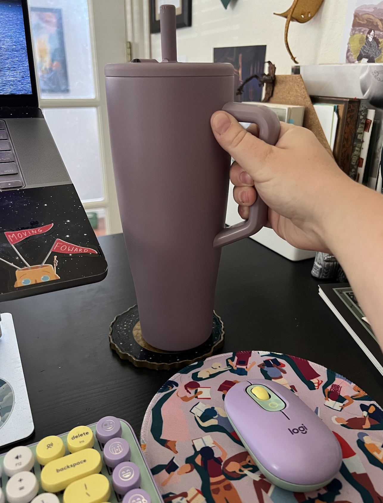 A manus  holding a ample  tumbler with a straw adjacent  to a laptop connected  a cluttered desk, featuring a colorful rodent  pad and keyboard
