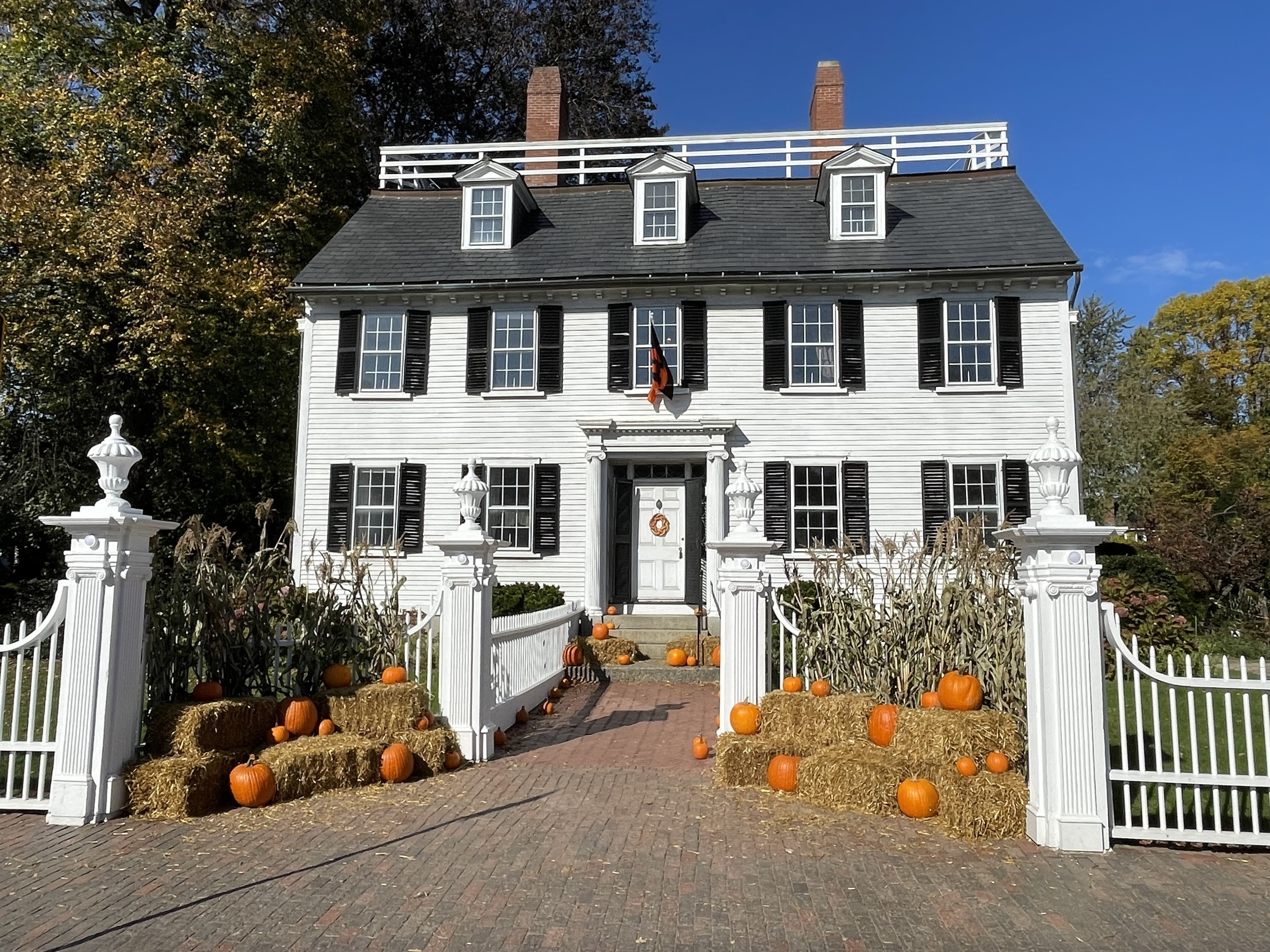 Historic two-story house decorated with pumpkins and hay bales, featuring a white picket fence and a brick pathway leading to the front door