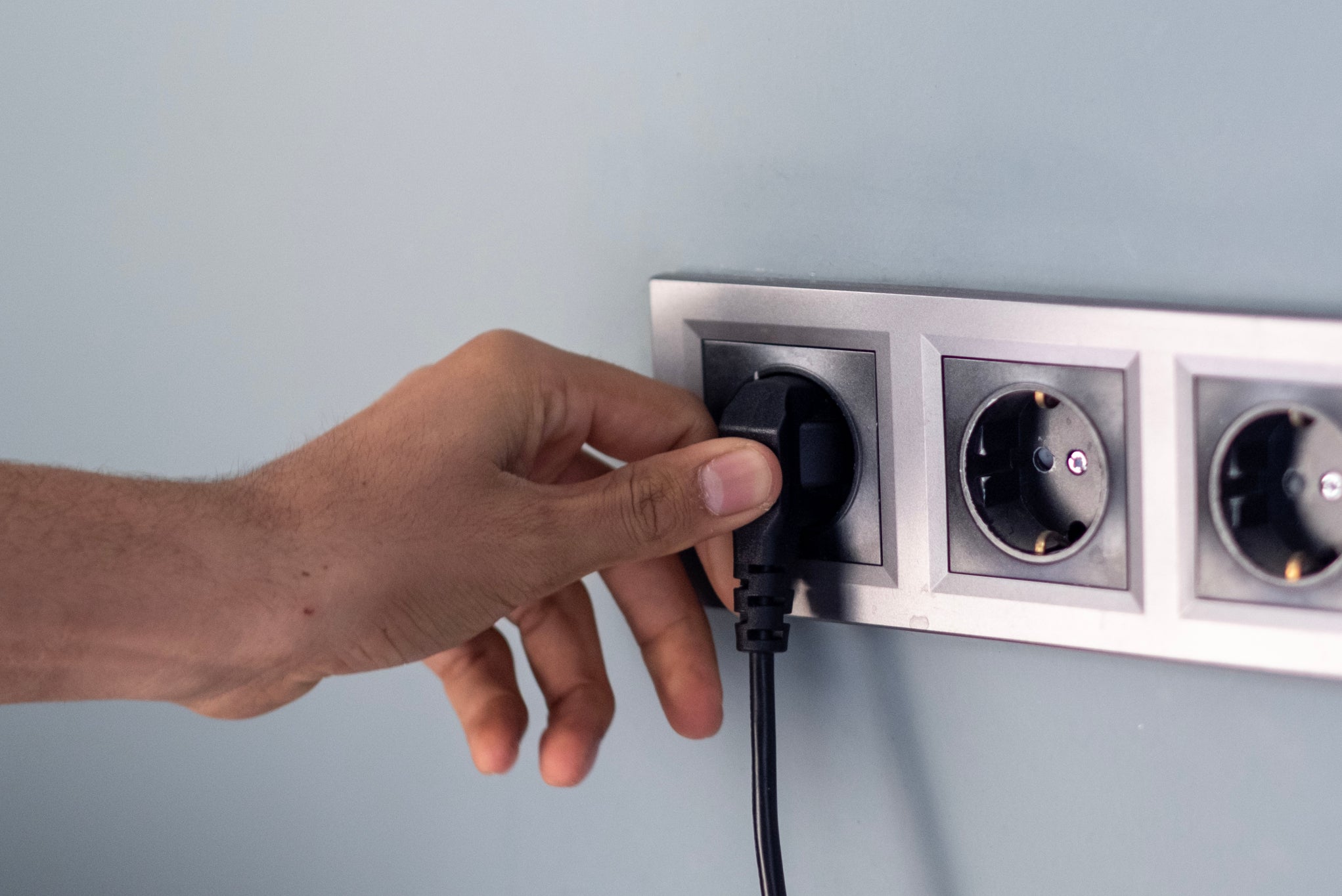 A hand is plugging a black cord into one of three electrical sockets on a wall