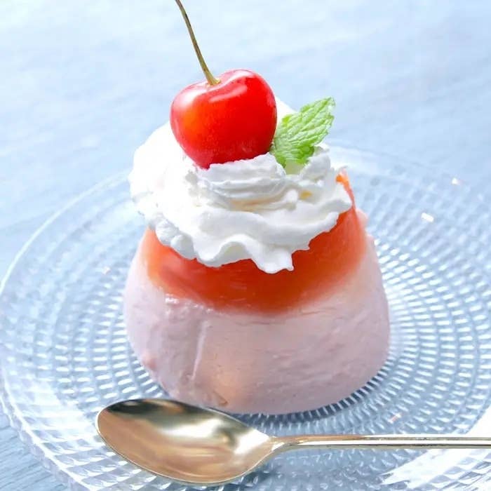 ガラスのプレートに乗ったピンク色のケーキ。ホイップクリームとサクランボ、ミントの葉が飾られ、側にスプーンが置かれている。