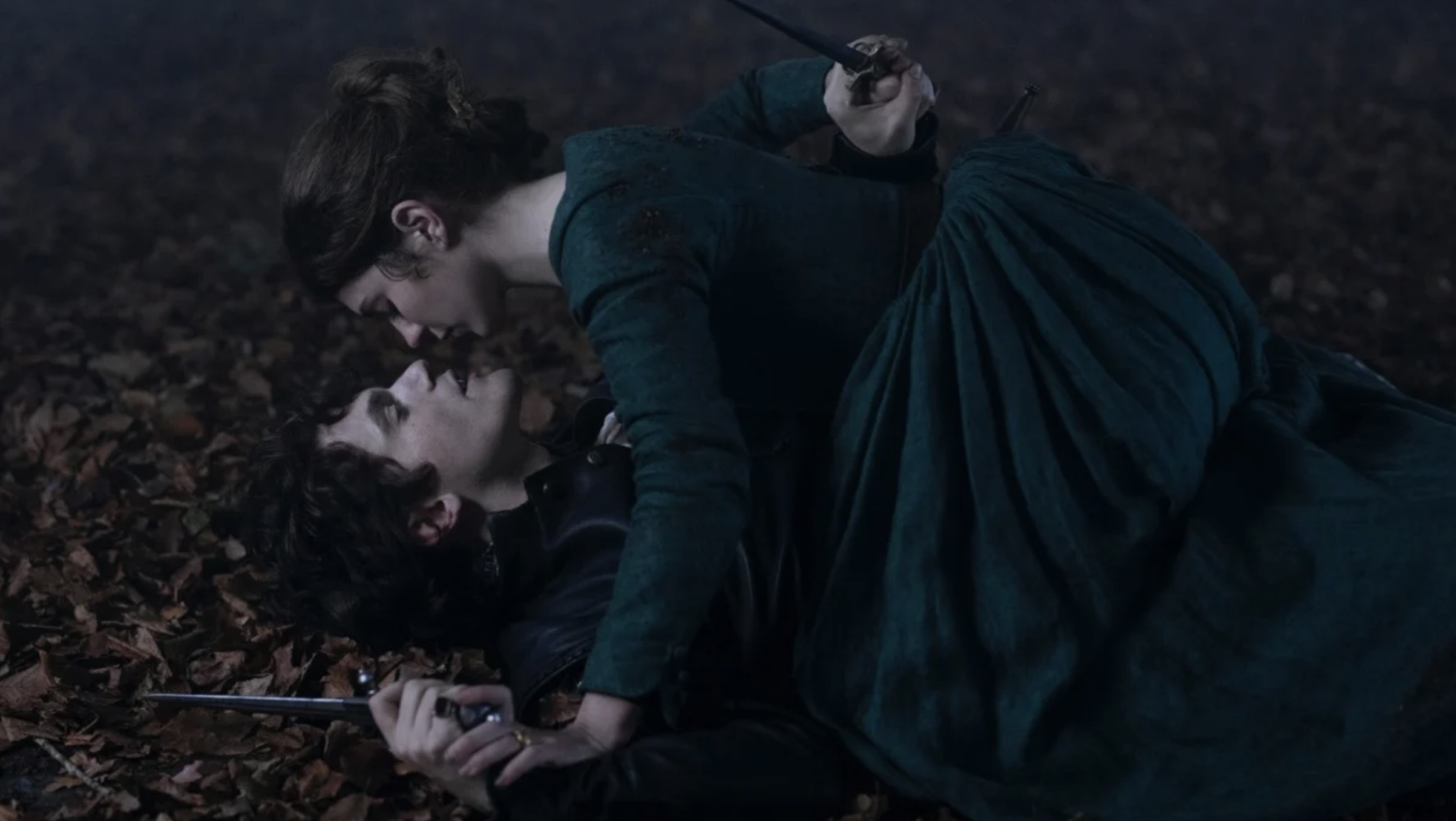 Dos personajes, uno encima del otro, sosteniendo pistolas y mirándose fijamente en el suelo cubierto de hojas