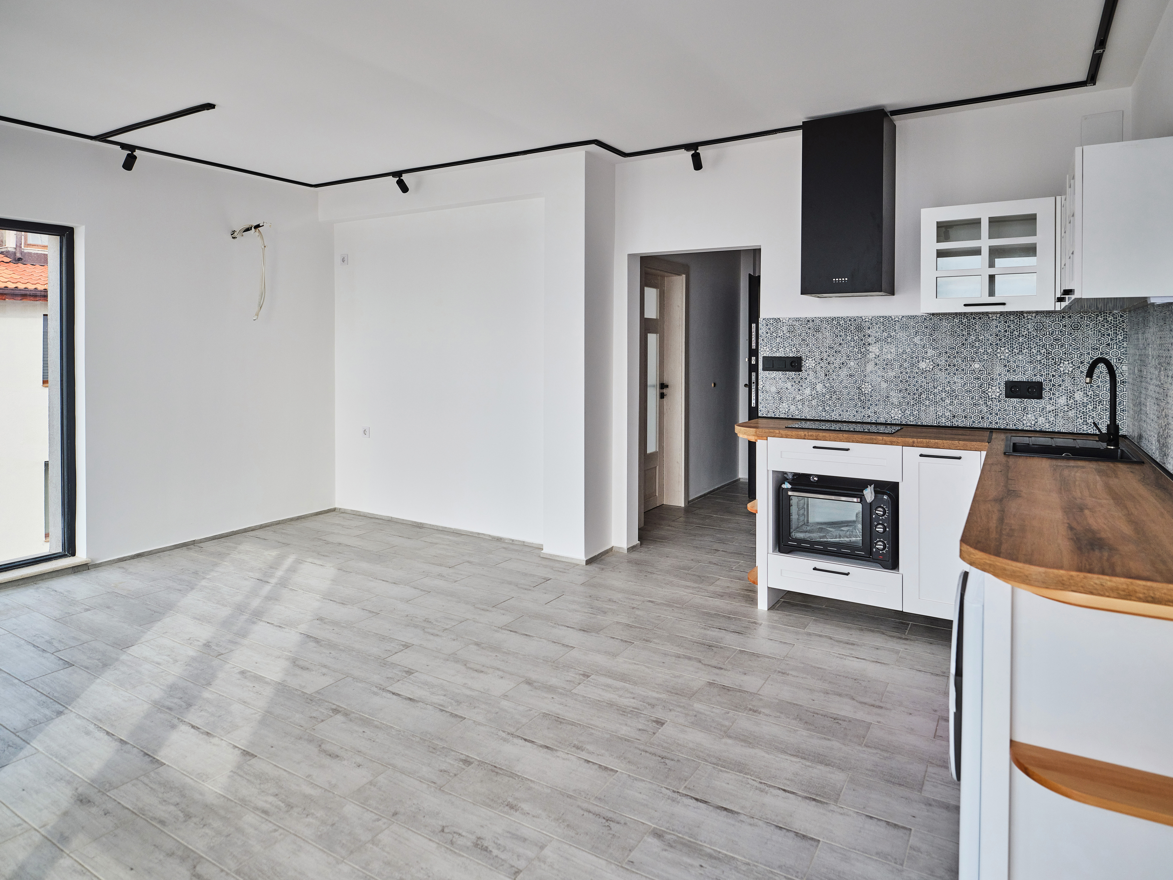 An empty modern kitchen with wooden countertops, an oven, and minimalistic cabinets adjacent to an open, spacious room with large windows