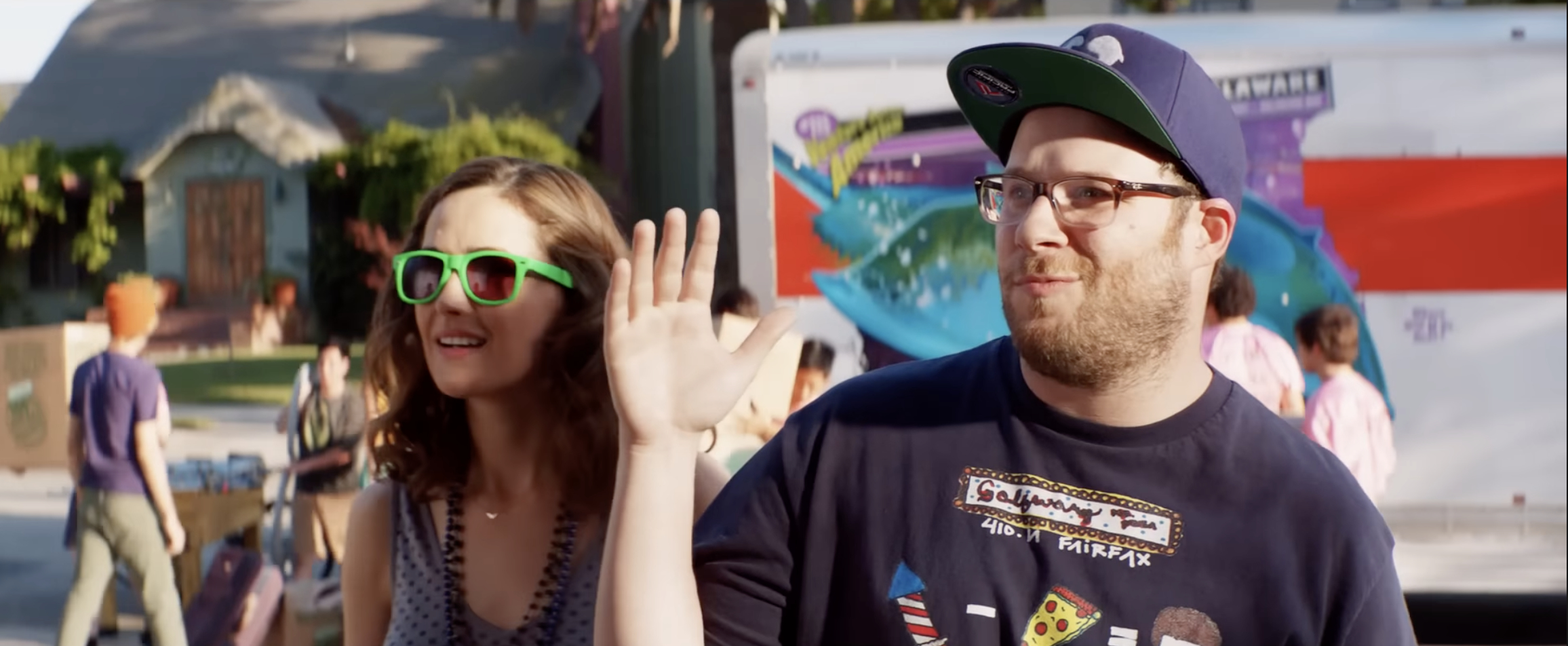 Rose Byrne and Seth Rogen smile and wave while standing outdoors. Seth wears a cap and glasses, and Rose wears sunglasses and a patterned top
