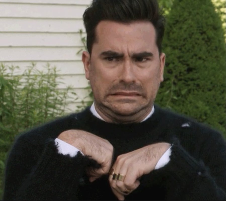 Dan Levy making a humorous grimace with hands up, wearing a black sweater on the set of "Schitt's Creek." Scene includes background greenery