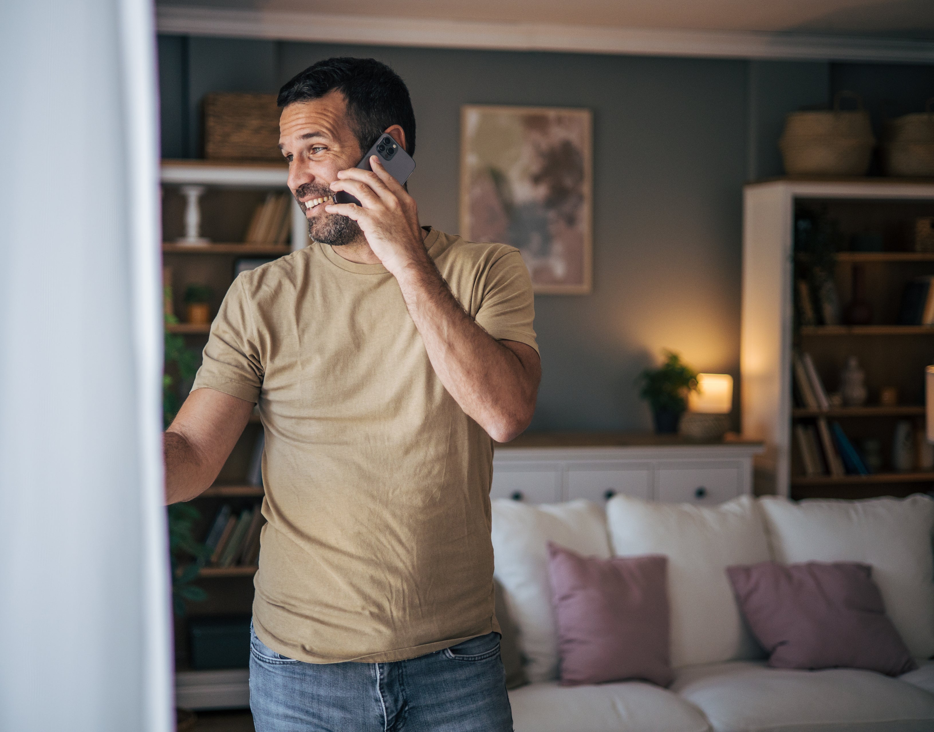 Man in a beige t-shirt talks on the phone in a cozy living room with shelves and a sofa. Names unknown