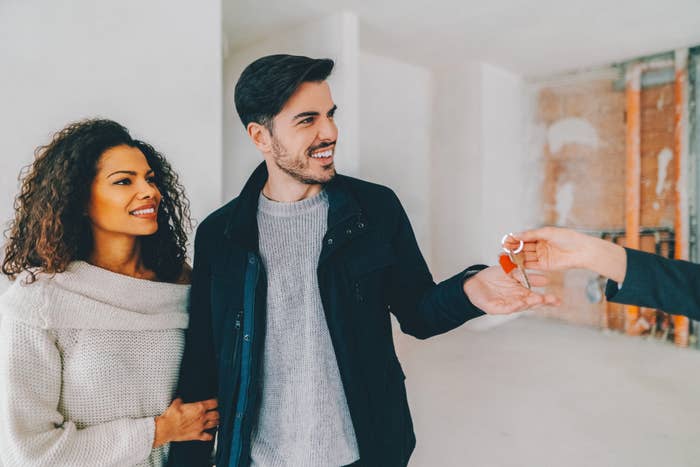 A man and woman happily accept keys to a new home from an unseen person. The couple wears casual, stylish clothing. The background shows an unfinished room