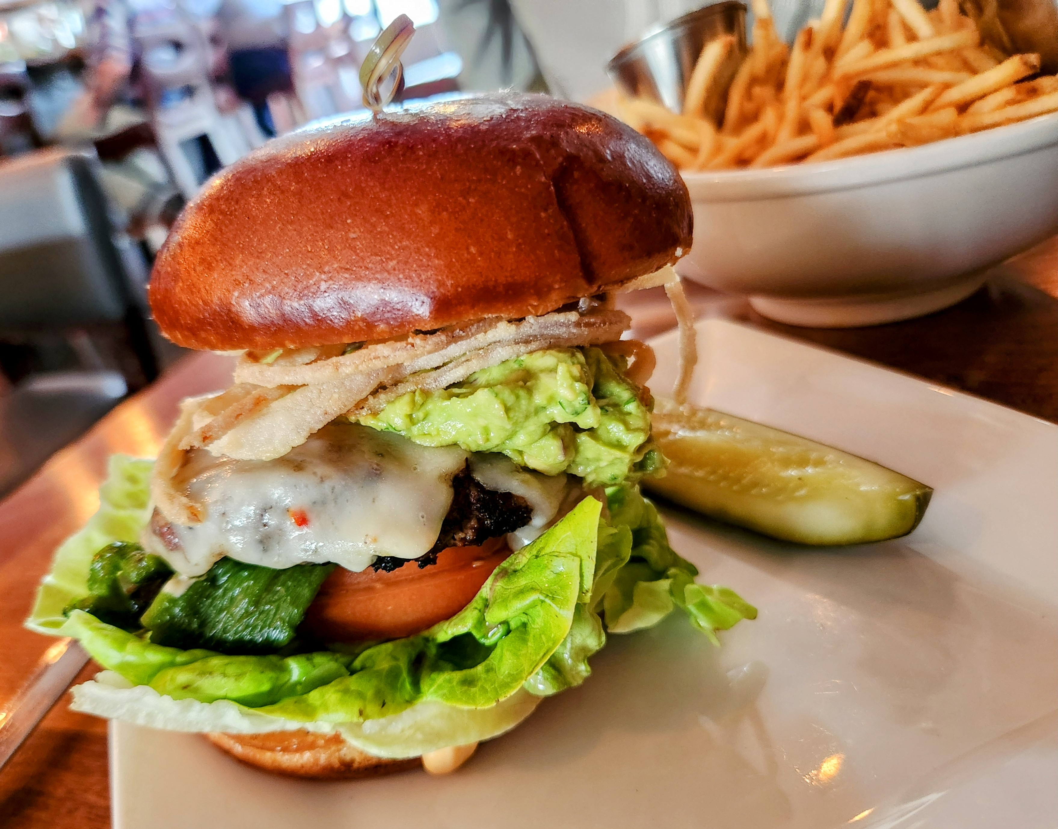 Close-up of a gourmet burger on a brioche bun, alongside a pickle and a bowl of fries in the background