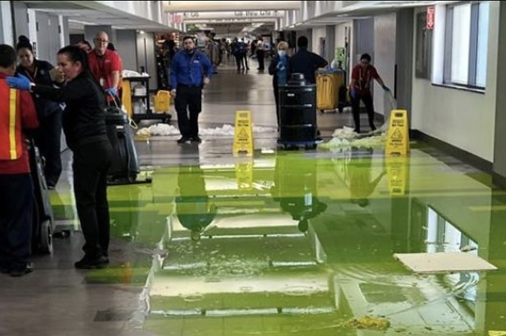 空港の通路が水たまりで溢れており、何人かの作業員が清掃している。複数の黄色い注意表示看板が置かれている。