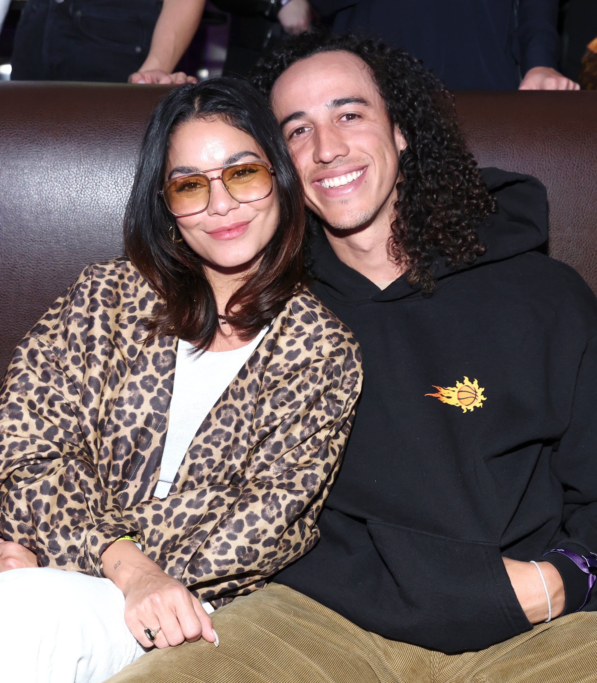 Vanessa Hudgens in a leopard-print coat and Cole Tucker in a black hoodie, sit together smiling on a couch