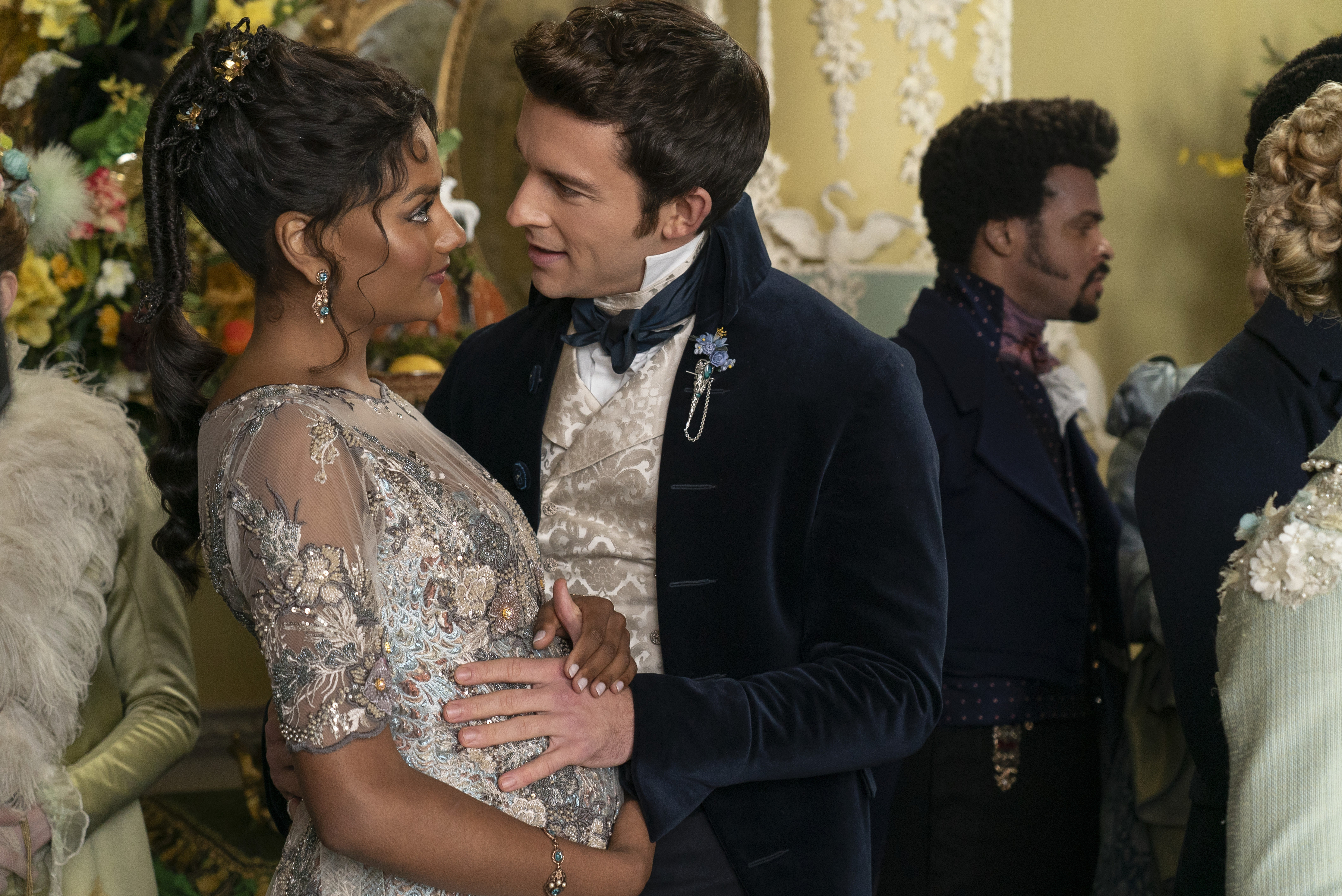 Simone Ashley and Jonathan Bailey, in period clothing, gaze at each other closely while at a formal event; other attendees are in the background