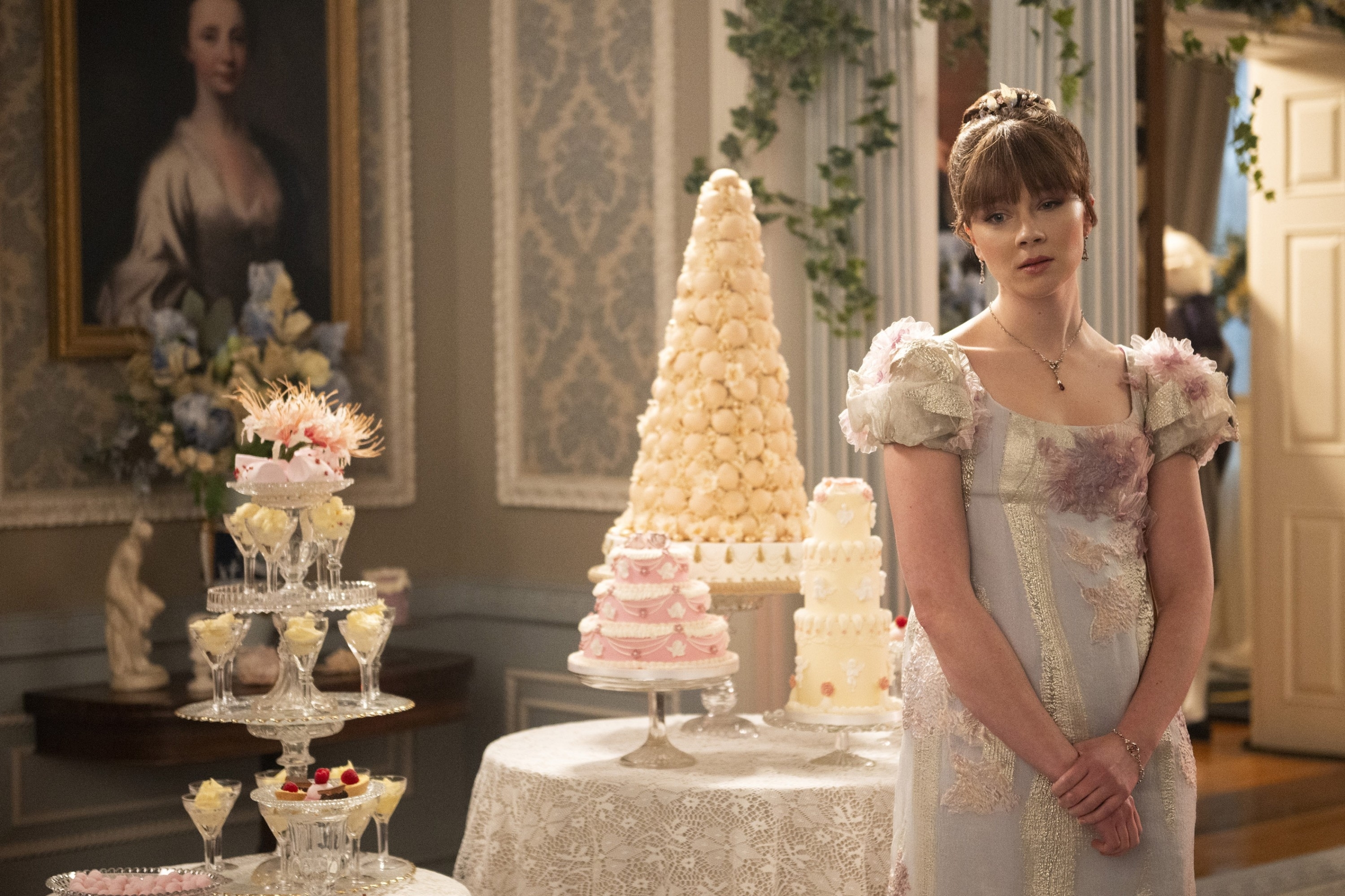 Claudia Jessie stands indoors in front of an elaborate dessert table wearing a period-style gown and diamond necklace