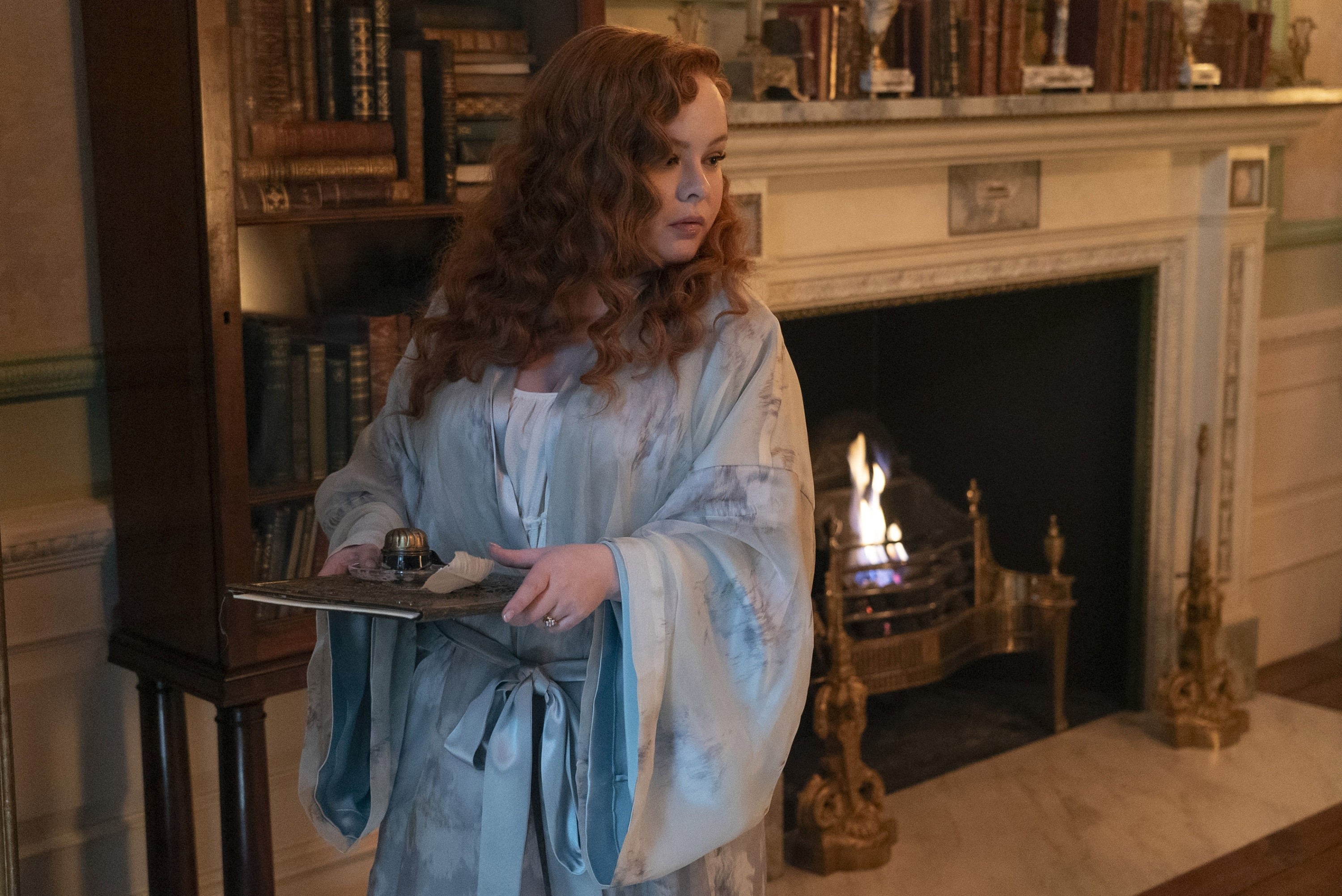Nicola Coughlan in a period costume robe stands by a fireplace, holding a book in one hand, looking to the side