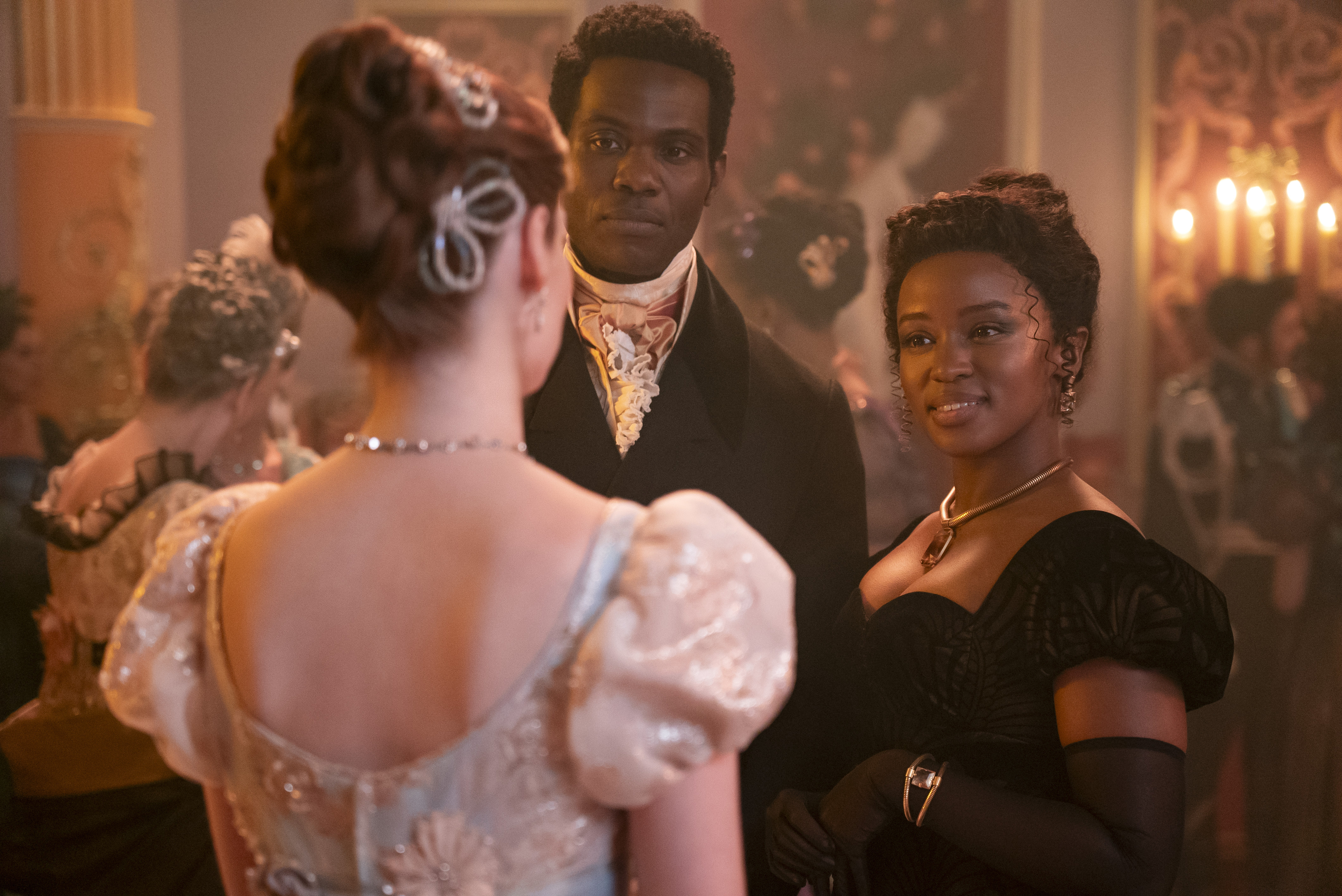 Three characters from Bridgerton, dressed in Regency-era formal attire, engage in conversation at a lavish party